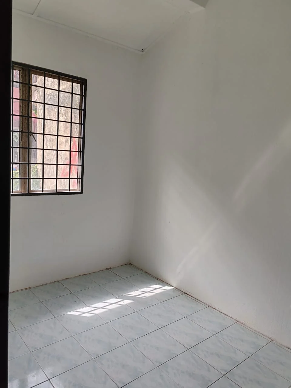 Room features: white painted walls, tile flooring, single window with metal grille, natural light from window.